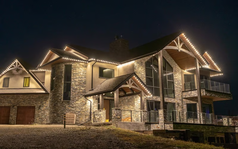 Timber-and-stone home outlined in warm white permanent lighting at night